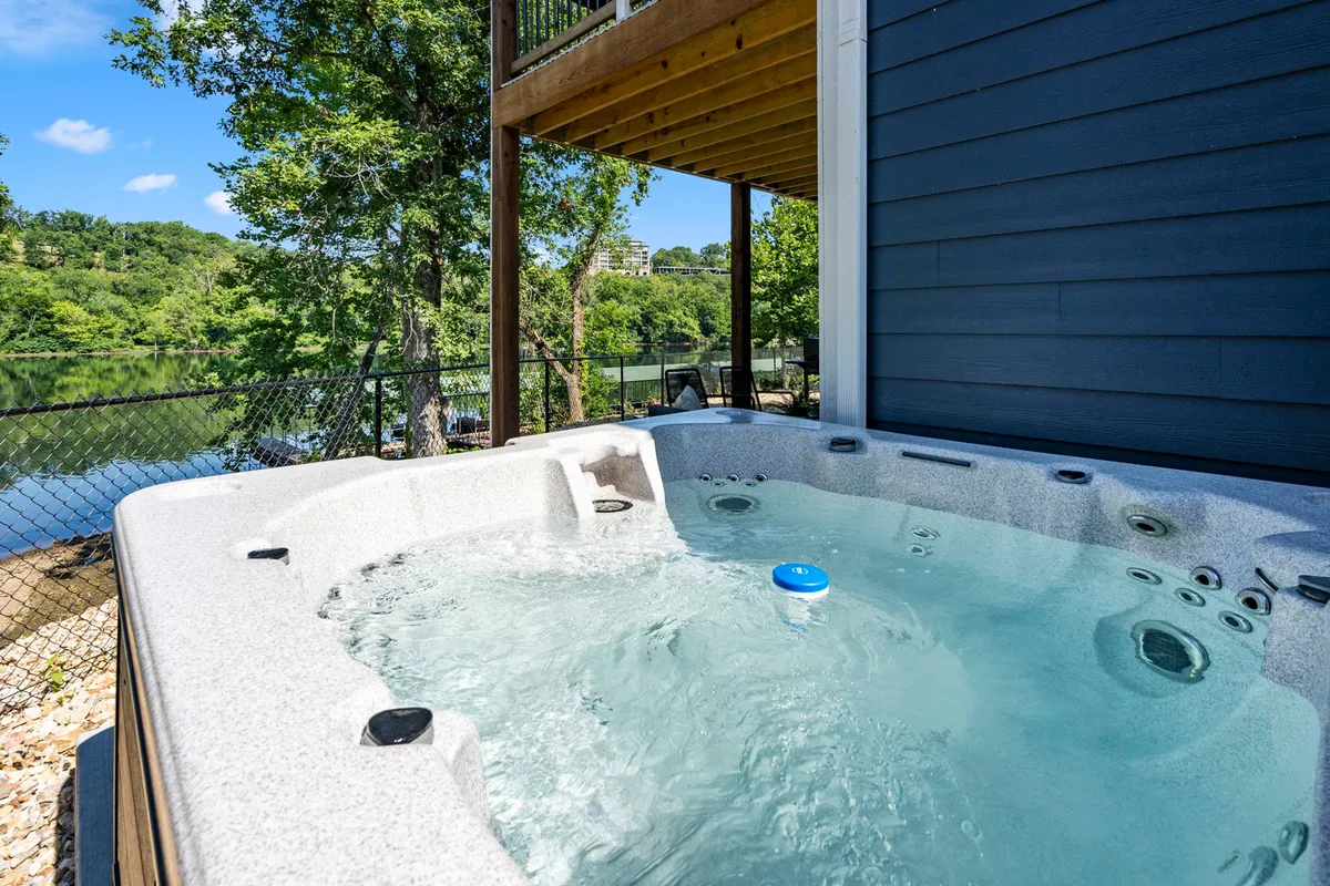 The private patio boasts a bubbling hot tub overlooking lush greenery and a shimmering water view. The space is perfect for unwinding while taking in the natural beauty of Lake Taneycomo, echoing the experience of relaxing outdoors with fresh air and scenic vistas.