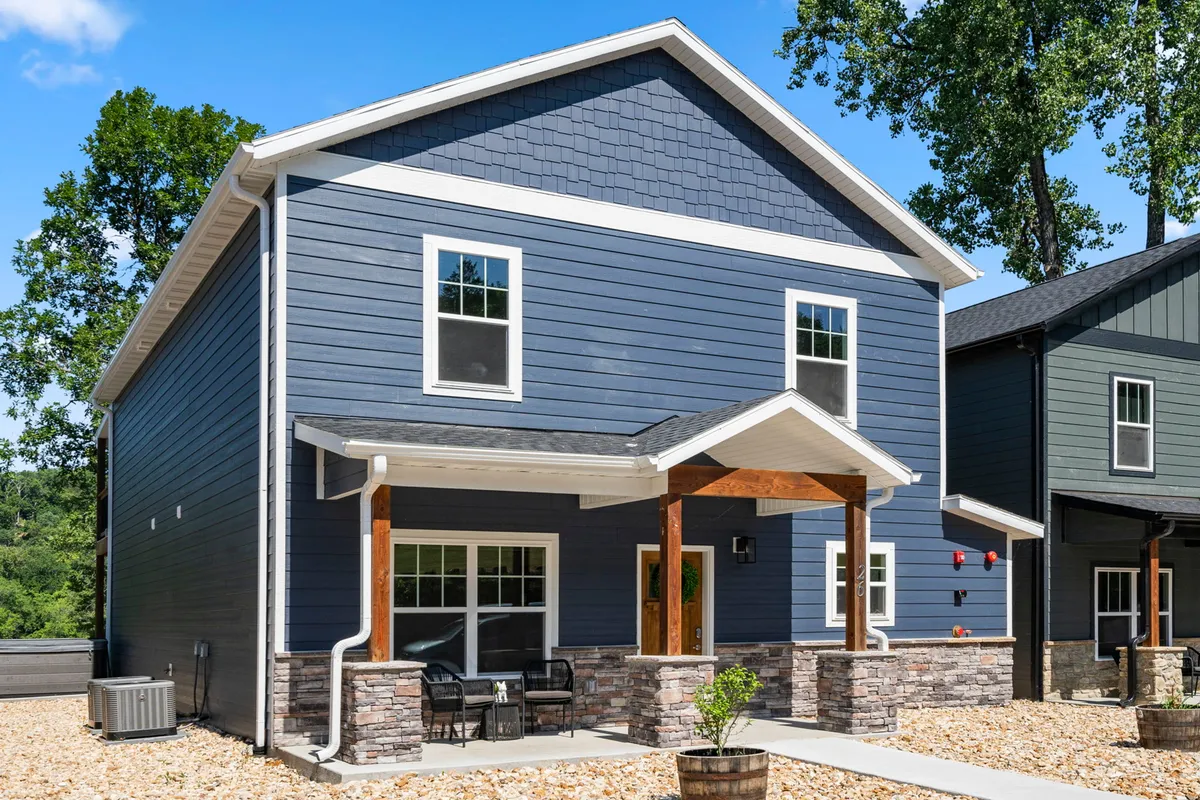 A cozy front porch with a stone foundation, blue siding, and large windows. The area features a welcoming vibe with space for socializing, relaxing, and enjoying neighborhood gatherings, capturing the spirit of community living.