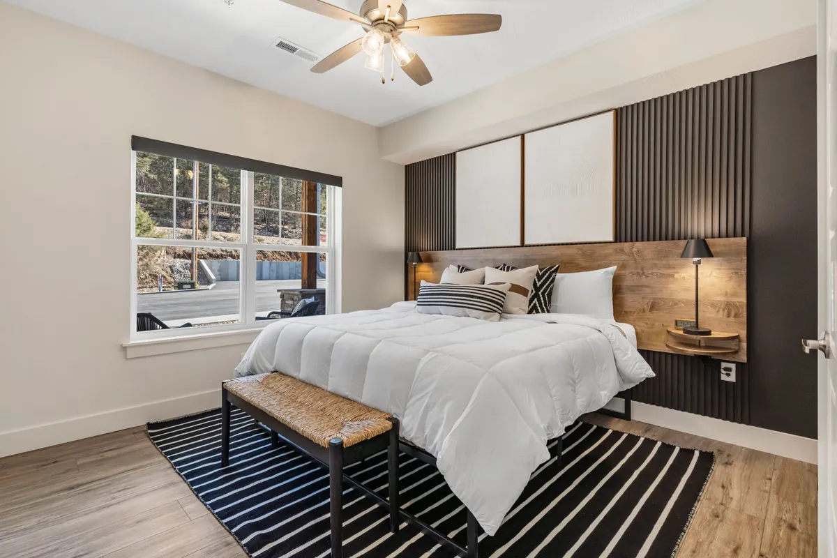 A cozy, light-filled king bedroom showcases a bed with decorative pillows, a striped rug, a window streaming in natural light, and tasteful wall art. The setting emphasizes comfort and a personal retreat for unwinding after lakeside adventures.