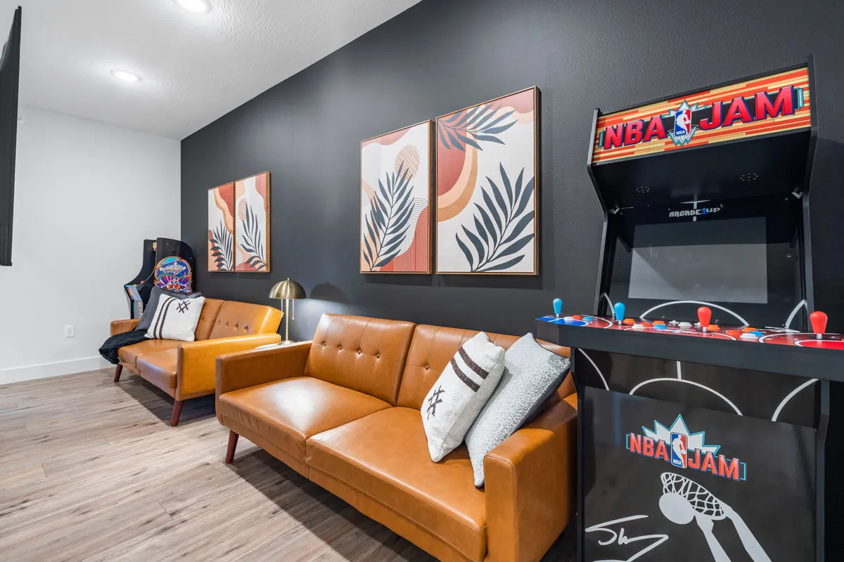 This interior scene showcases a classic NBA Jam arcade machine, brown leather sofas, and vibrant tropical wall art. The casual setup is clearly designed for gaming, socializing, and relaxation, embodying a youthful, upbeat atmosphere.