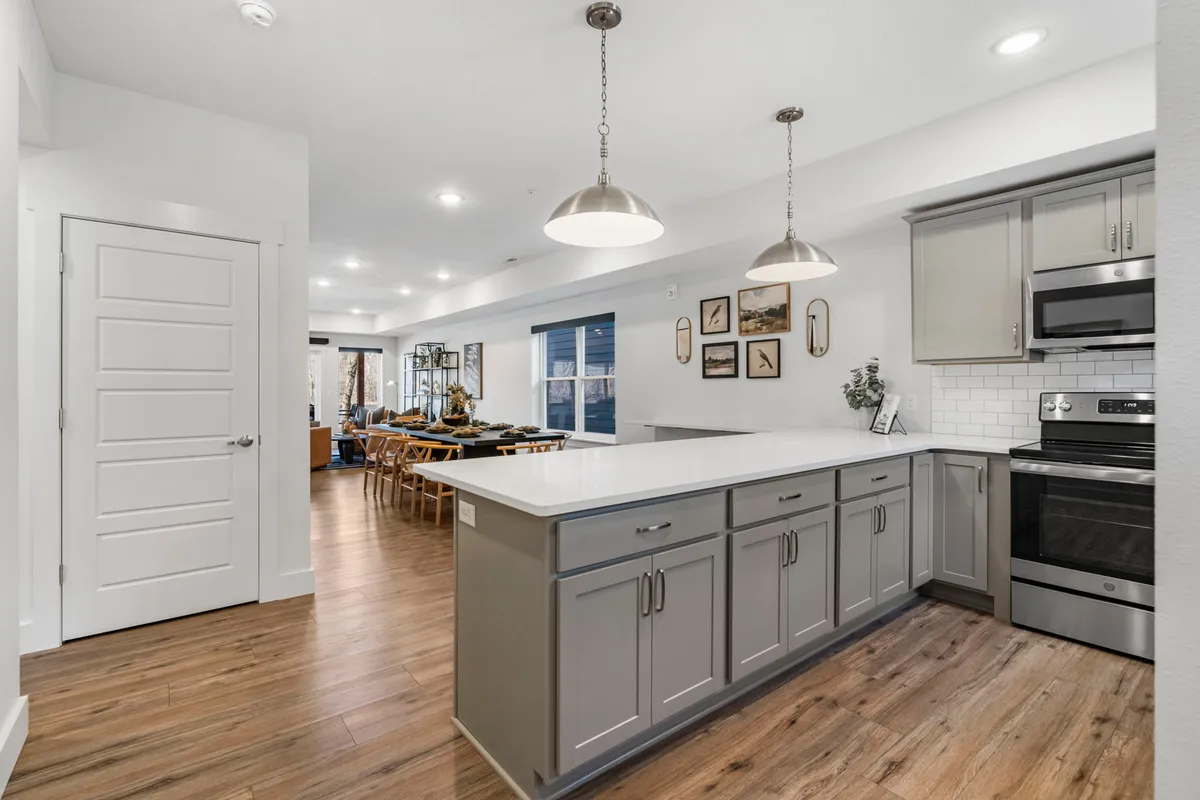 An open-plan kitchen and dining area featuring a central kitchen island with stainless steel appliances, bar stools for casual seating, and a spacious dining table positioned nearby. Natural light streams in, highlighting the modern yet inviting atmosphere perfect for group gatherings and shared meals.