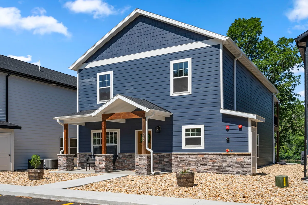 A welcoming exterior shot of a blue house with stone accents, a covered porch, and large windows. The porch area is set up for socializing and relaxation, offering a glimpse into the inviting community atmosphere.
