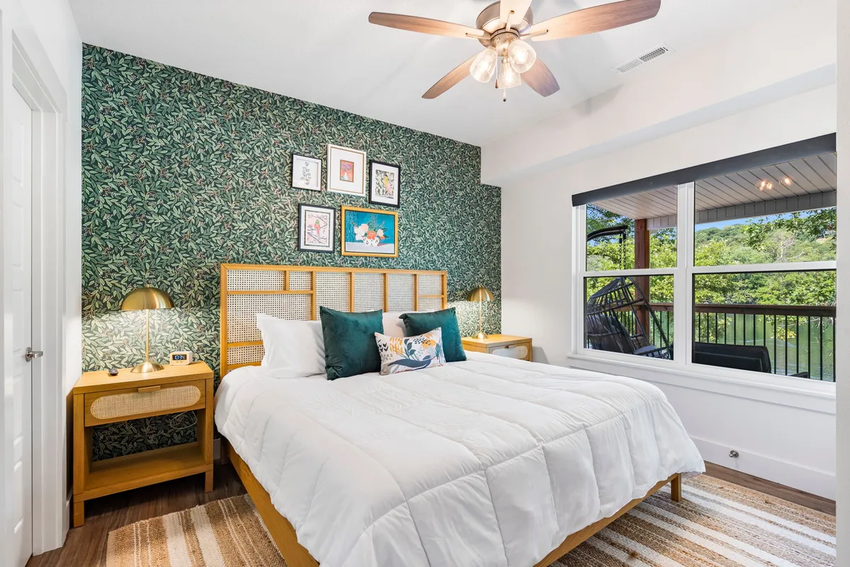 A bright and refreshing king bedroom features a bed with crisp white bedding, a soothing green accent wall, framed artwork, nightstands with lamps, and a large window offering a tranquil view of the outdoors. The room exudes calmness and comfort, ideal for restful nights and sunny mornings.
